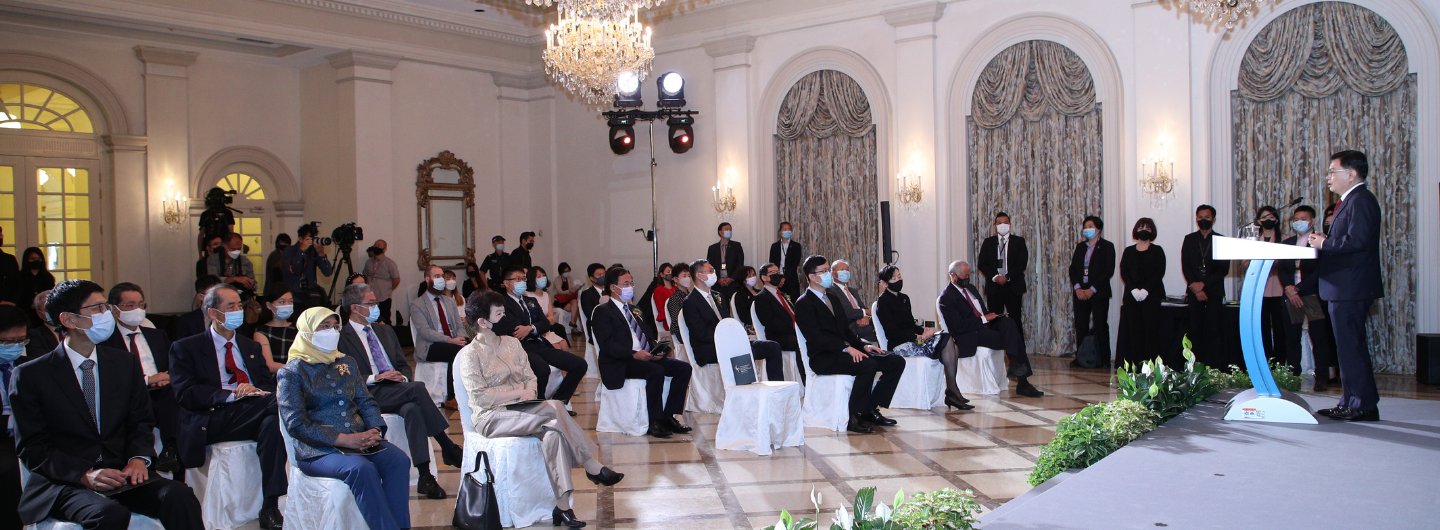 Speaker on stage addresses seated audience; many wear face masks in ornate hall.